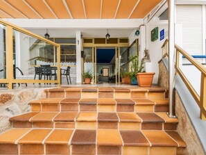 Un escalier extérieur mène à un espace abrité où se trouvent une table, des chaises, des plantes en pot et une statue.