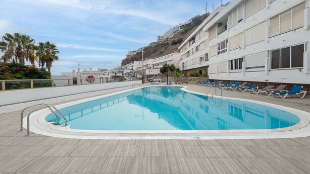 A swimming pool with a wooden deck, lounge chairs, and a building with balconies overlooking a rocky hillside.