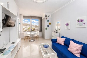 A modern living room with a blue sofa, a round dining table with chairs, a flat-screen TV mounted on the wall, and a large window offering a view of buildings and hills. - Sun Port Ocean Views - Renovated & Pool 2 (Puerto Rico)