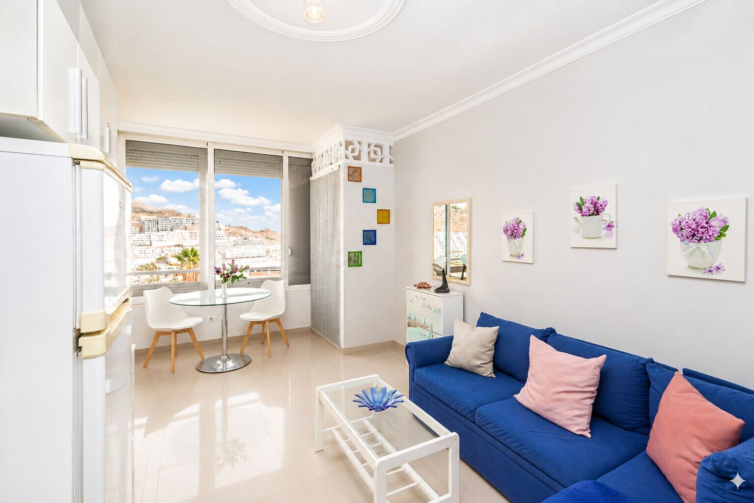 A modern living room with a blue sofa, a glass coffee table, and a dining area with a round table and chairs.