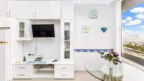 A modern room with a flat-screen TV mounted on a white cabinet, a glass table with a vase of flowers, and a view of a cityscape through a window. - Sun Port Ocean Views - Renovated & Pool 2 (Puerto Rico)