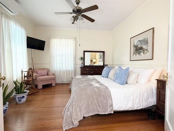 Ein Schlafzimmer mit einem Bett, einem Deckenventilator, einer Kommode, einem Fernseher und einem Fenster mit Vorhängen.