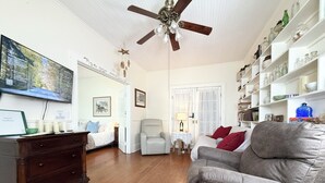 Un salon avec un plancher en bois, un ventilateur de plafond, un téléviseur, une bibliothèque et un fauteuil.