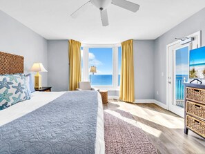 Un dormitorio con cama, mesita de noche, lámpara, ventana con cortinas, ventilador de techo y vista al océano.