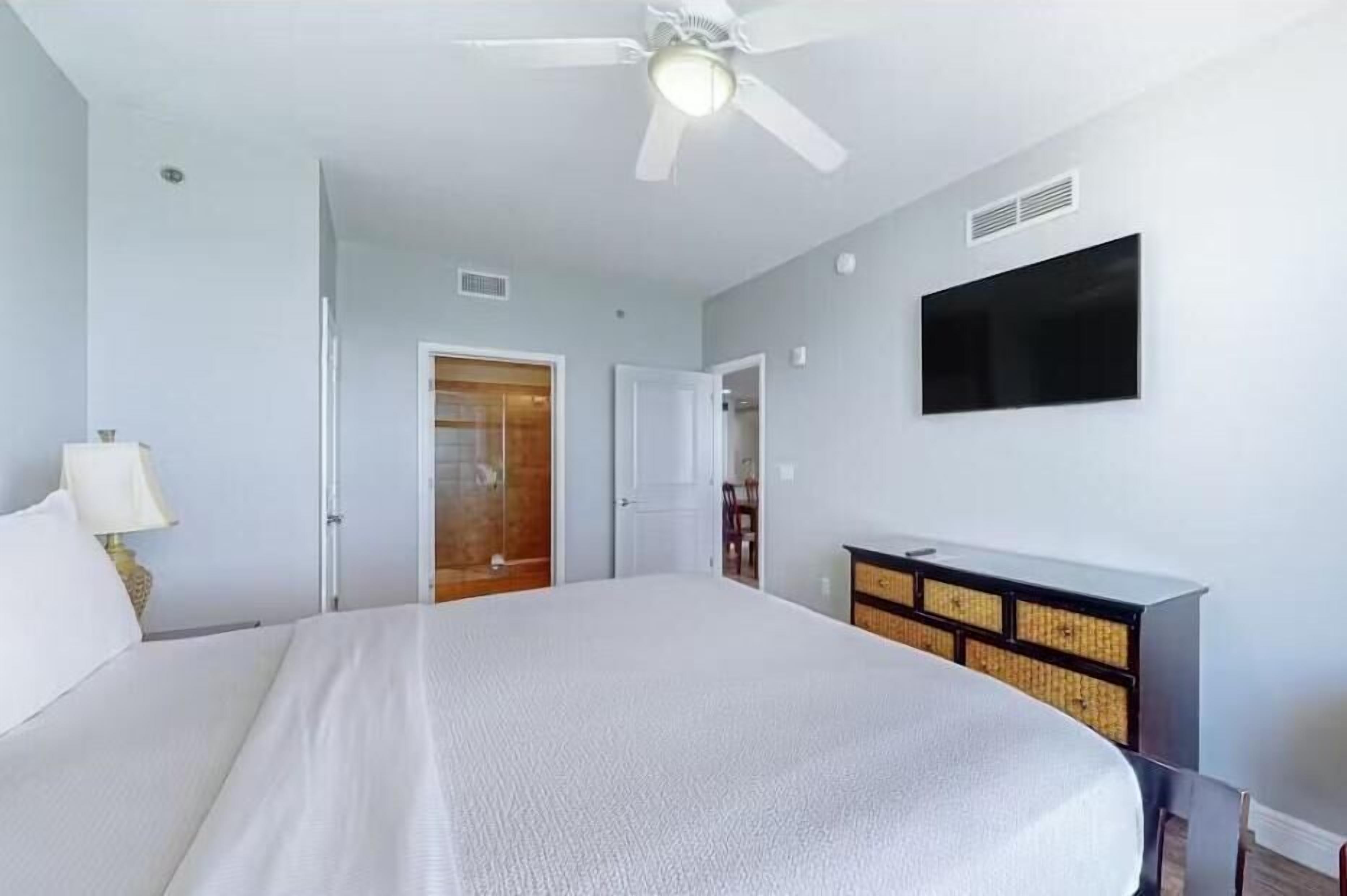 Ein Schlafzimmer mit einem Bett, einem Deckenventilator, einem an der Wand montierten Fernseher und einer Holztür.