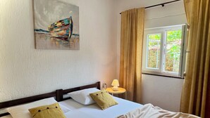 Ein Schlafzimmer mit Bett, Kissen, einer Nachttischlampe, einem Fenster mit Vorhängen und einem Bild an der Wand.