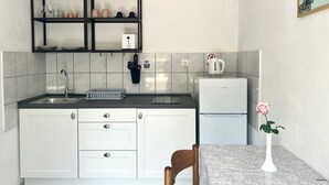 Eine kleine Küche mit weißer Arbeitsplatte, einem schwarzen Hängeschrank darüber und einem kleinen, weißen Kühlschrank.