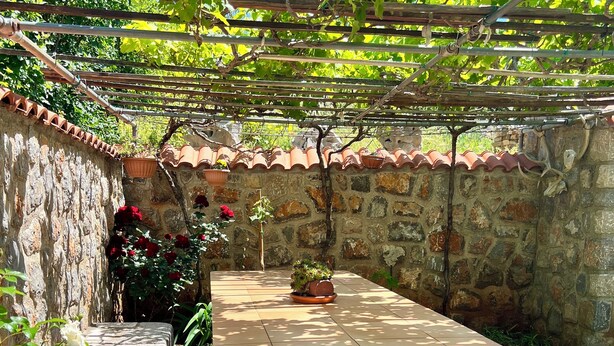 Eine Steinterrasse mit Tisch und Stühlen, umgeben von einer Steinmauer und einem mit Kletterpflanzen bewachsenen Pergola.