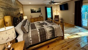 A bedroom with a wooden floor, a bed with a forest-themed bedspread, a nightstand with a lamp, a dresser, a chair, a TV mounted on the wall, and a window with curtains. - Tranquil Escape: Hot Tub, Scenic Views, Family Fun (Ranger)