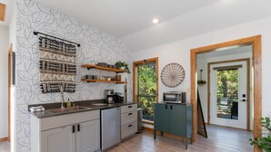 A modern kitchen with a wooden floor, built-in cabinets, a stainless steel sink, and a microwave on a cabinet. There are shelves with plants and kitchen items, and a large window with a view of greenery. - King Bed | Boho Charm | 2 minutes to Downtown (Eureka Springs)