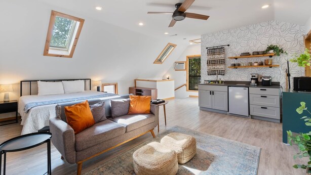 A modern, well-lit room with a bed, sofa, ottomans, and a small table. There is a ceiling fan, a skylight, and a kitchen area with cabinets and a countertop. - King Bed | Boho Charm | 2 minutes to Downtown (Eureka Springs)