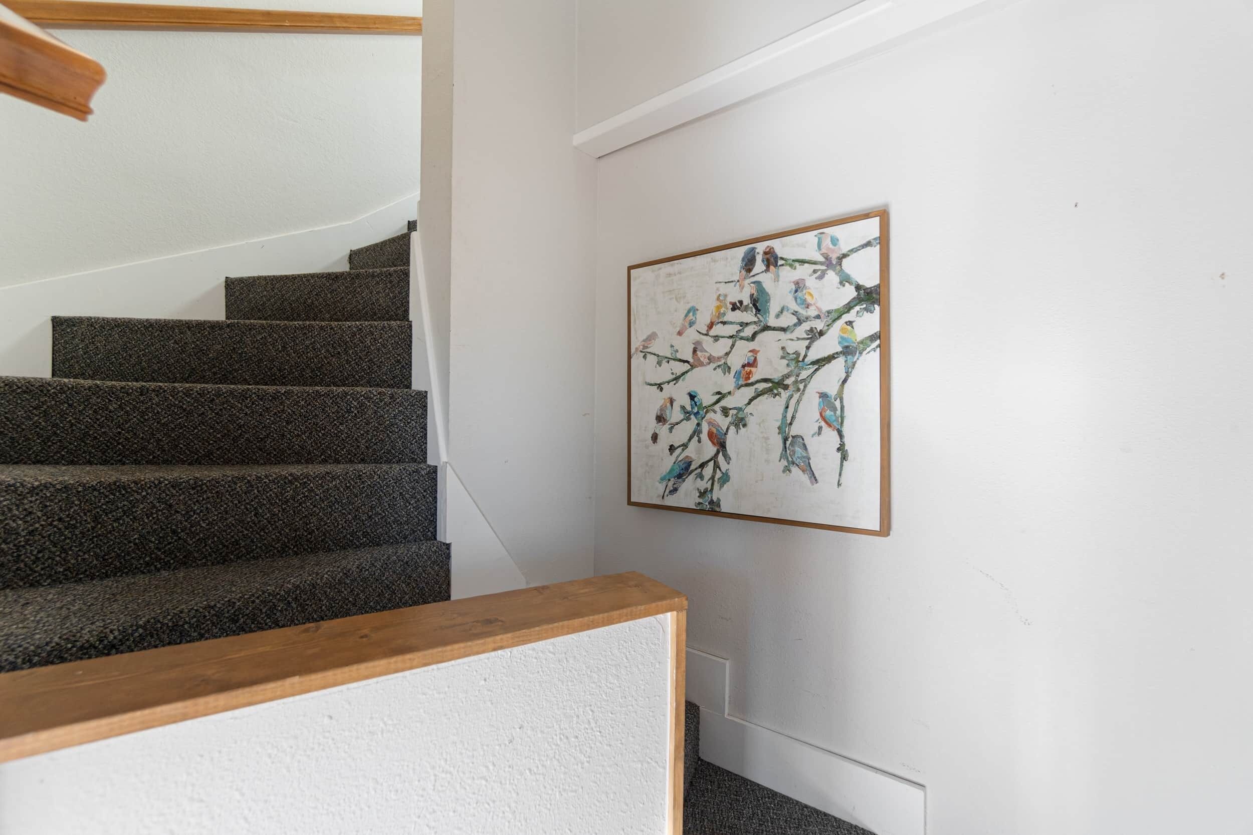 A staircase with dark steps and a wooden handrail, leading to a white wall with a framed painting of birds on branches.
