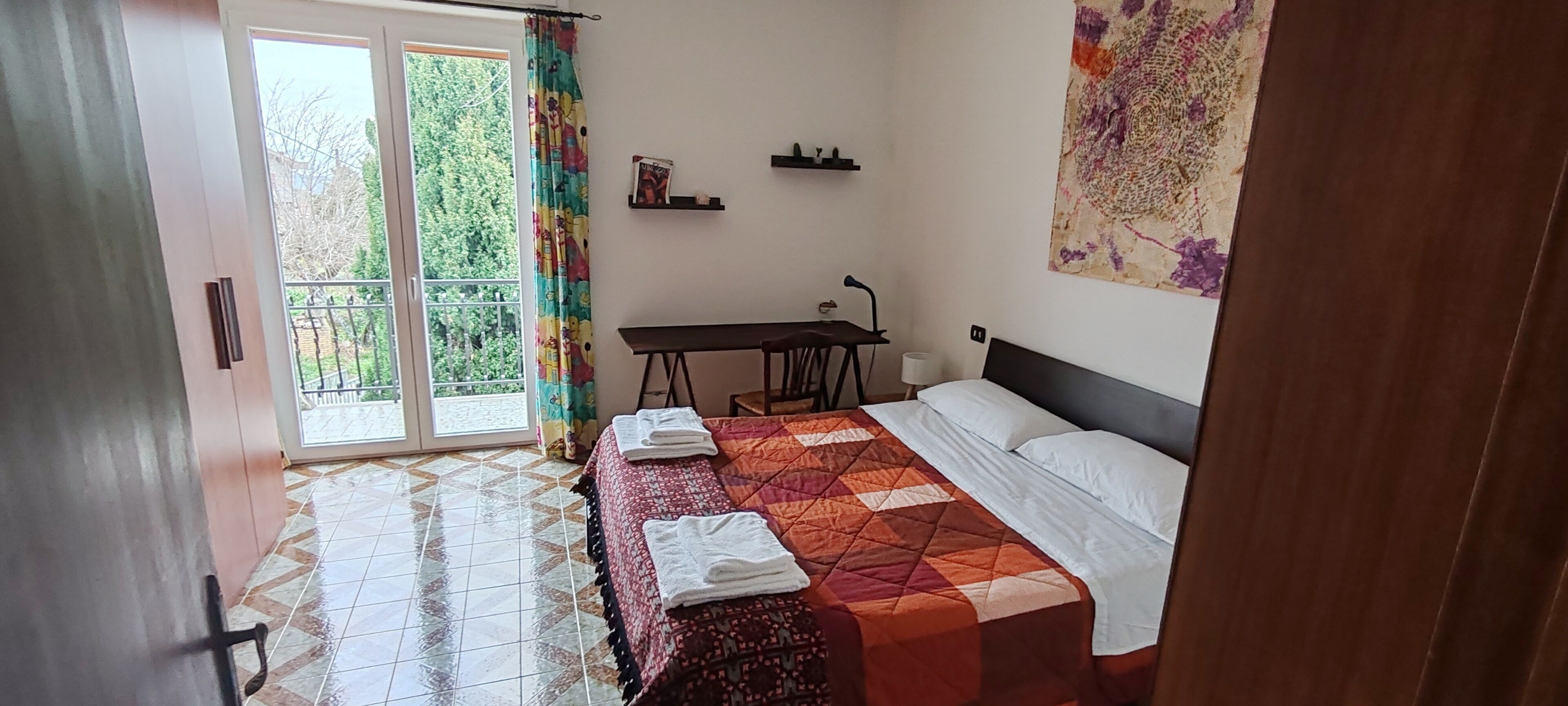 Un dormitorio con una cama, un escritorio y una silla. Hay una ventana con cortina, un estante en la pared y un cuadro.