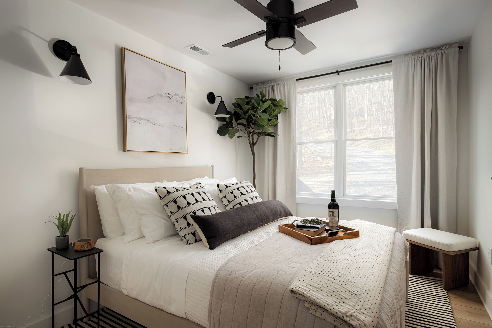 Une chambre à coucher comprenant un lit, une table de chevet, une lampe, un ventilateur de plafond, une fenêtre, des rideaux et une plante.