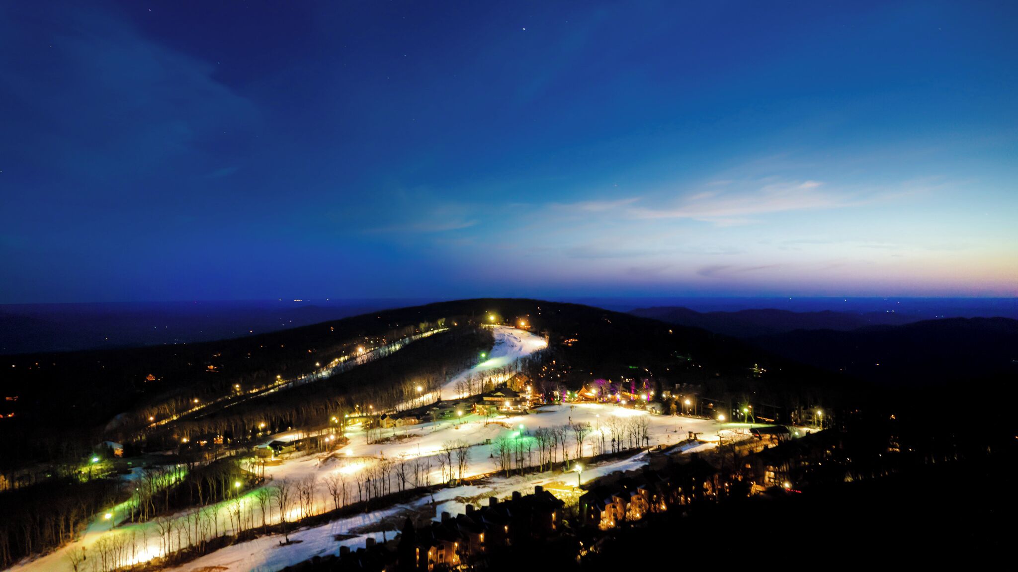 A snow-covered landscape with a winding road and illuminated buildings, likely a ski resort at night.