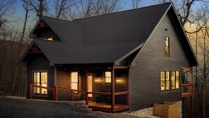 Une cabane en rondins à deux étages, avec une façade de couleur gris foncé, une terrasse en bois et des fondations en pierre.