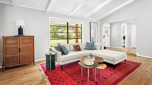 A spacious living room with a large sectional sofa, a coffee table, and a red patterned rug. There is a wooden cabinet with a lamp on top, and a window with a view of greenery outside. - Luxurious & Tranquil 3-bedroom House & Garden in Houston (Houston)