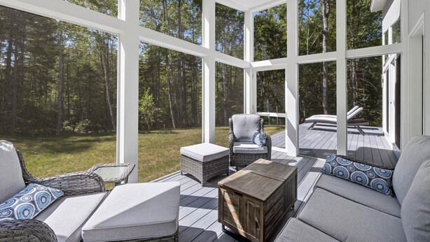 A covered outdoor seating area with wicker furniture, a wooden coffee table, and a view of a deck and forest. - Summer Breeze at Goose Rocks Beach (Kennebunkport)