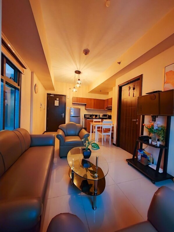 A modern living room with leather sofas, a glass coffee table, and a dining area with wooden cabinets and a kitchen in the background.
