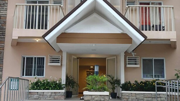 Entrada de um hotel com vários andares, com acesso central ladeado por duas varandas, degraus de pedra e plantas em vasos.