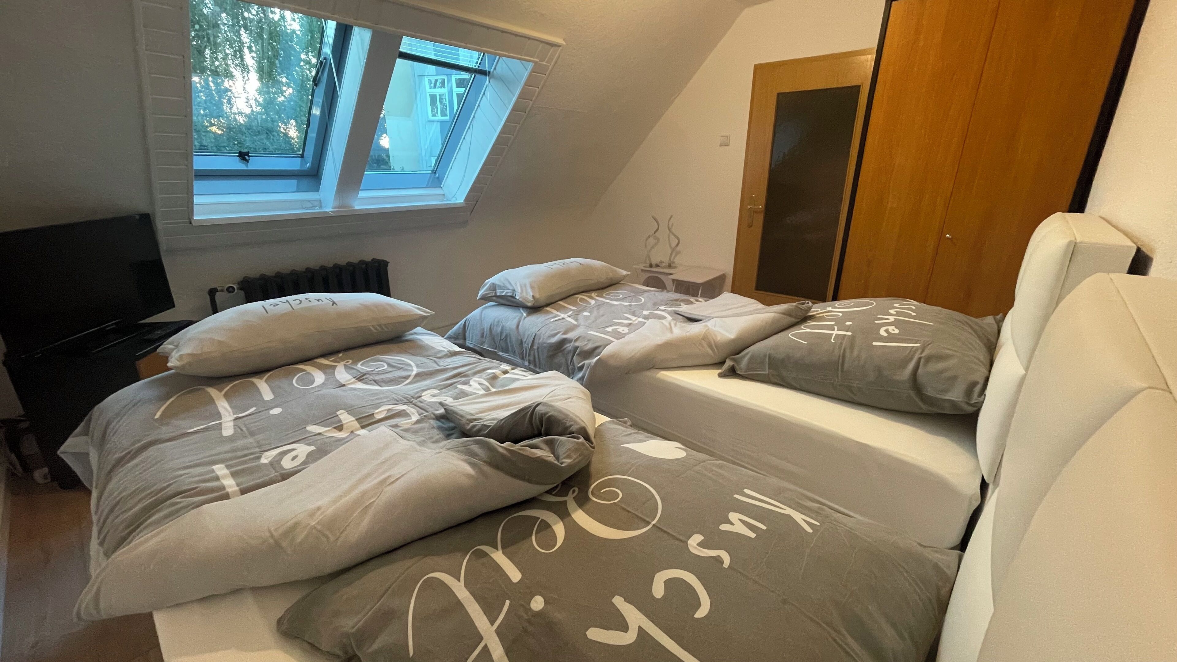 A bedroom with a sloped ceiling, a skylight, a bed with grey bedding, a wooden door, and a radiator.