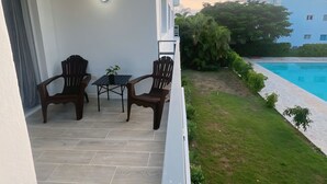 Varanda com duas cadeiras de madeira e uma pequena mesa, uma parede branca e vista para uma piscina e vegetação.