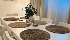 Uma área de refeições com mesa branca, jogos americanos trançados, cadeiras brancas, uma planta em vaso e um relógio de parede.