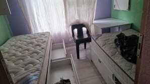 A small, compact room with two beds, a chair, and a desk.