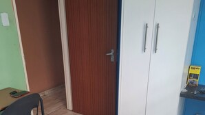 A room with a wooden door marked with the number 6, a white wardrobe with two handles, a black plastic chair, and a bed with a patterned bedspread.