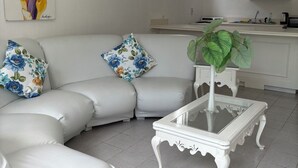 A modern living room with a white sofa, floral cushions, a glass coffee table, and a kitchen area in the background. - Runaway Bay Caldo Tours (Runaway Bay)