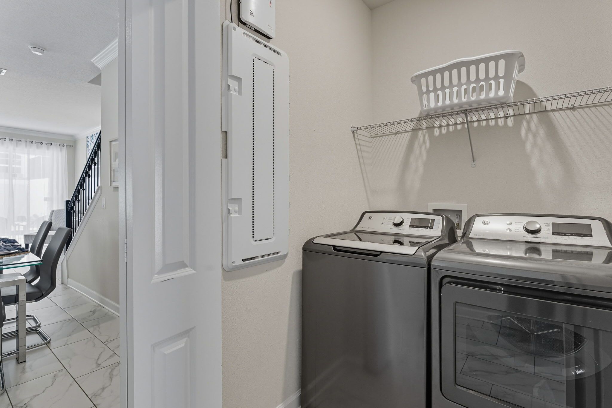 A laundry room with two washing machines and a dryer, a white wall-mounted laundry basket, and a staircase leading to another room.