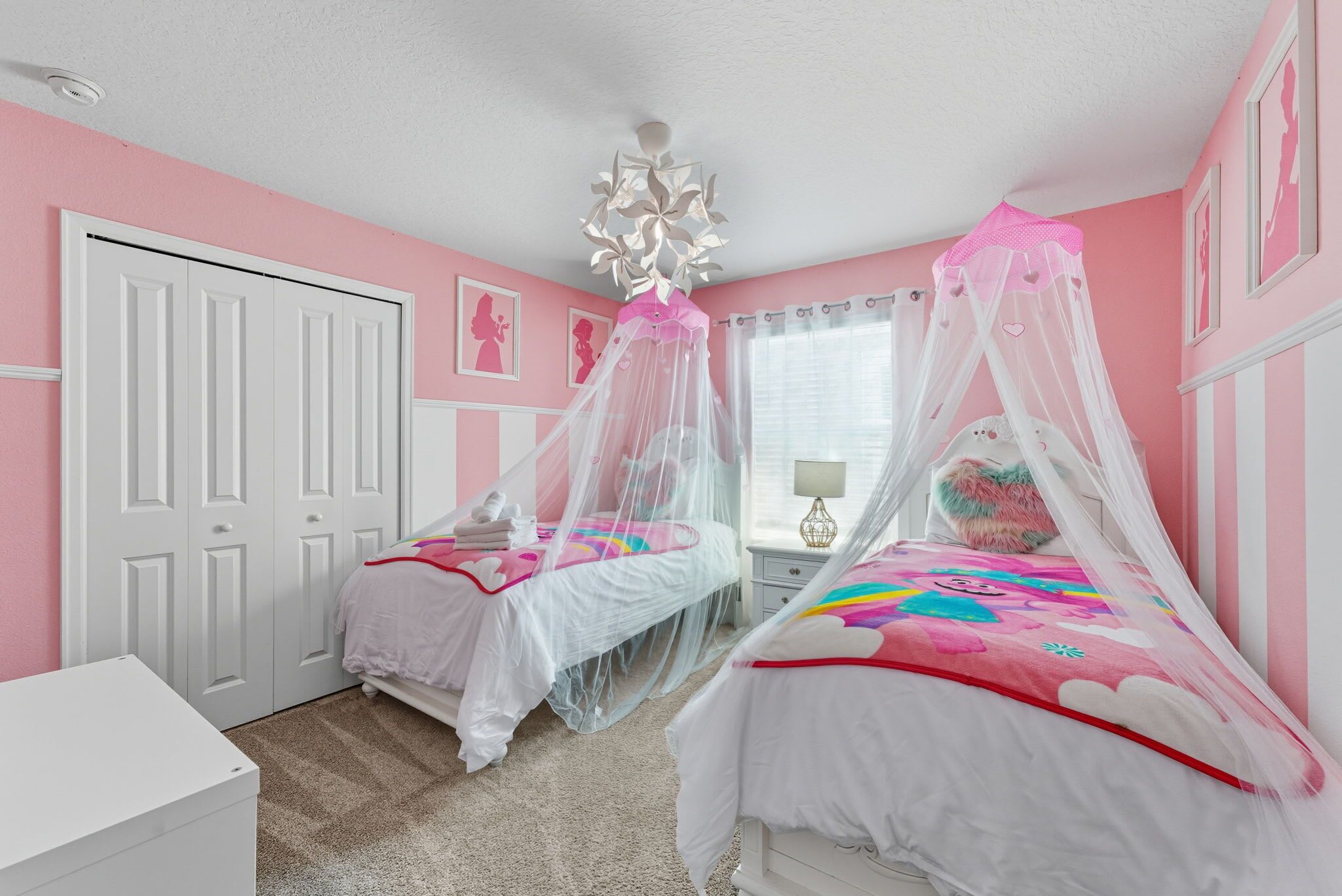A pink-themed bedroom with two beds, each with a canopy, a white dresser, and a ceiling light fixture.