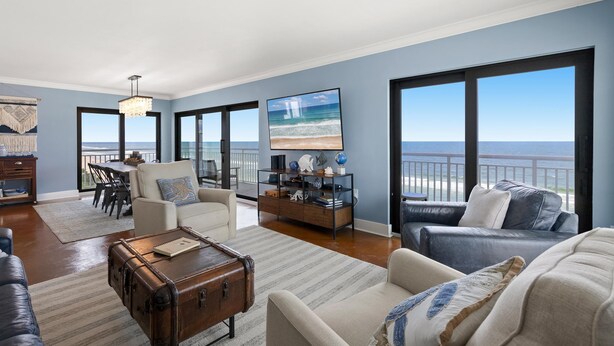 A spacious living room with a large window overlooking the beach, a cozy seating area, and a dining space. - Shorehom by the Sea 75 (New Smyrna Beach)