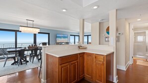 A modern kitchen with a large island, wooden cabinets, and a view of the ocean through large windows. - Shorehom by the Sea 75 (New Smyrna Beach)