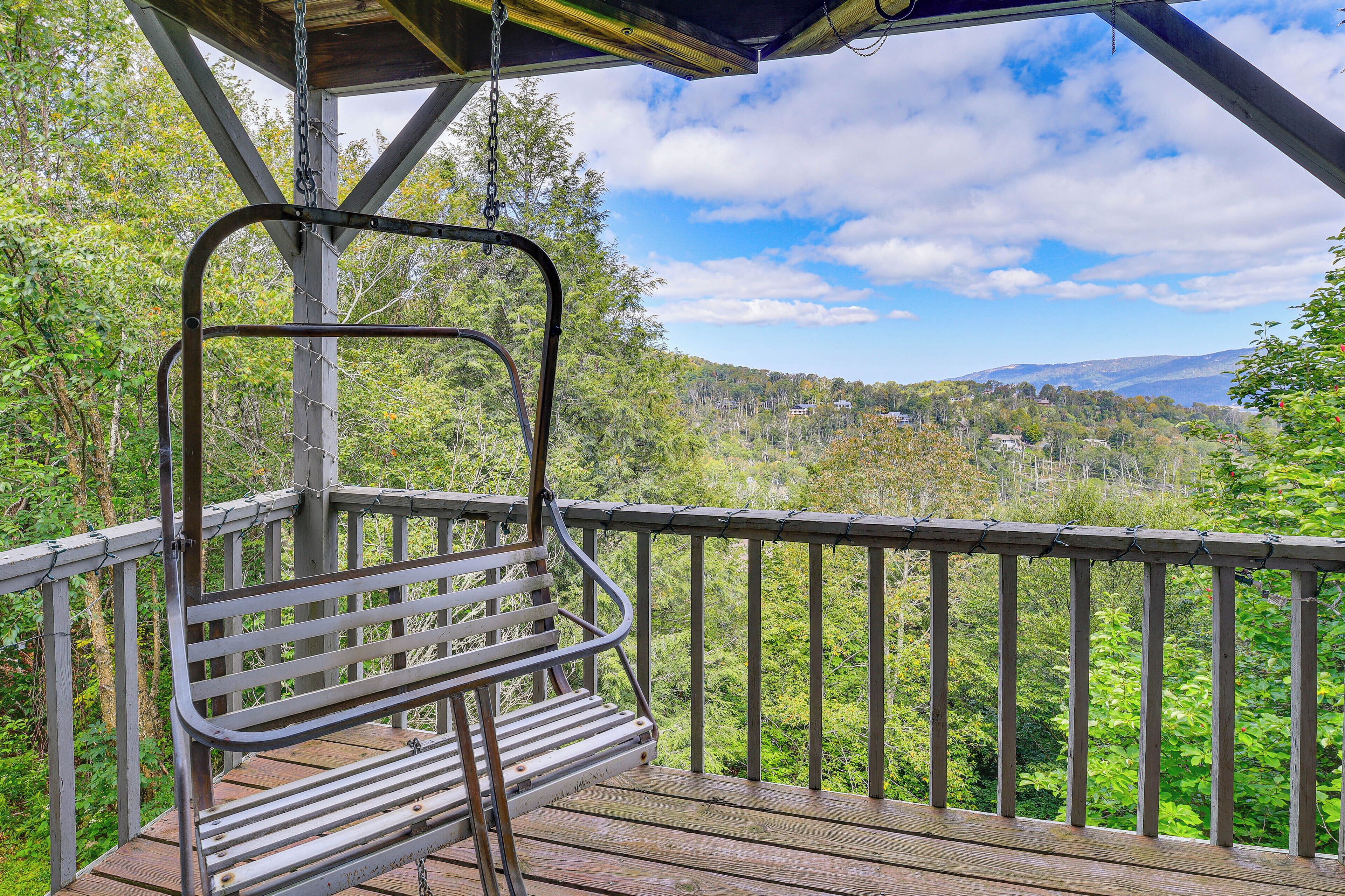 Une terrasse en bois avec un banc et une rambarde en métal, offrant une vue sur une vallée boisée et des montagnes lointaines.