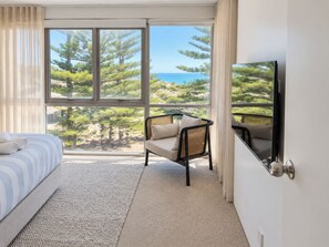 A hotel room with a bed, a chair, a large window with a view of trees and the sea, and a television on the wall. - Shoreline Retreat - beach frontage - 2 King Beds (Scarborough)