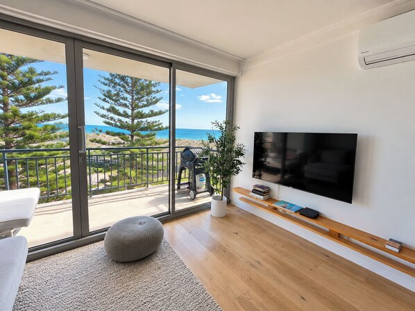 A modern living room with a large TV, wooden flooring, a balcony with a view of the sea, and a potted plant.