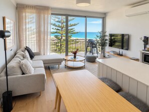 A modern living room with a sofa, coffee table, and TV, overlooking a balcony with a view of the sea and trees. - Shoreline Retreat - beach frontage - 2 King Beds (Scarborough)
