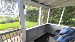 Un porche abrité avec une chaise en osier et des coussins, un paillasson noir et une vue sur une cour verdoyante et des arbres.
