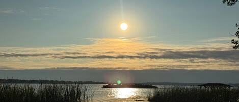 Ein Sonnenuntergang über ruhigem Wasser mit Reflexionen der Sonne, umgeben von hohem Gras und einer fernen Küste.