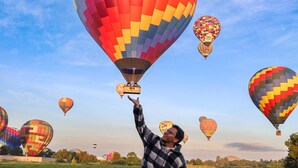 Una persona señala un globo aerostático colorido en un campo, con varios globos en el fondo.