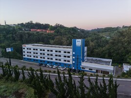 Un hotel de varios pisos con un llamativo esquema de colores azul y blanco, un estacionamiento con varios autos, y un fondo de vegetación exuberante y colinas.