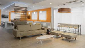 Una sala moderna con sofá, otoman y mesa de centro, un área de cocina con taburetes altos y un ventanal con persianas.