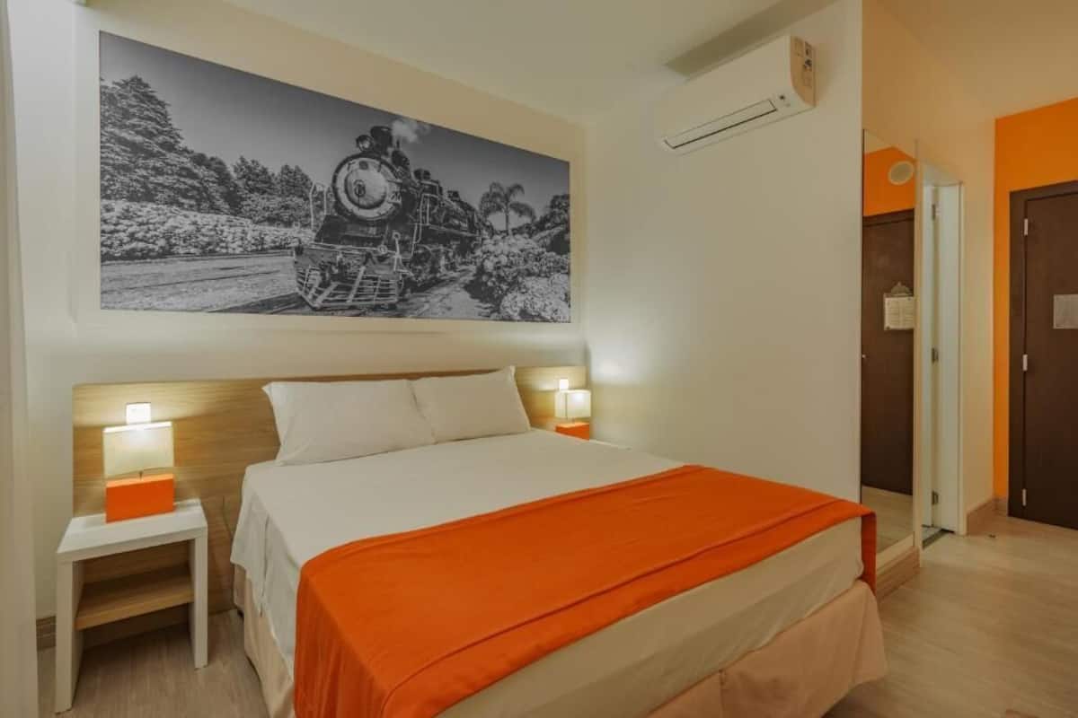 Una habitación de hotel con una cama grande, dos mesitas de noche con lámparas, un aire acondicionado de pared y una puerta a la derecha.