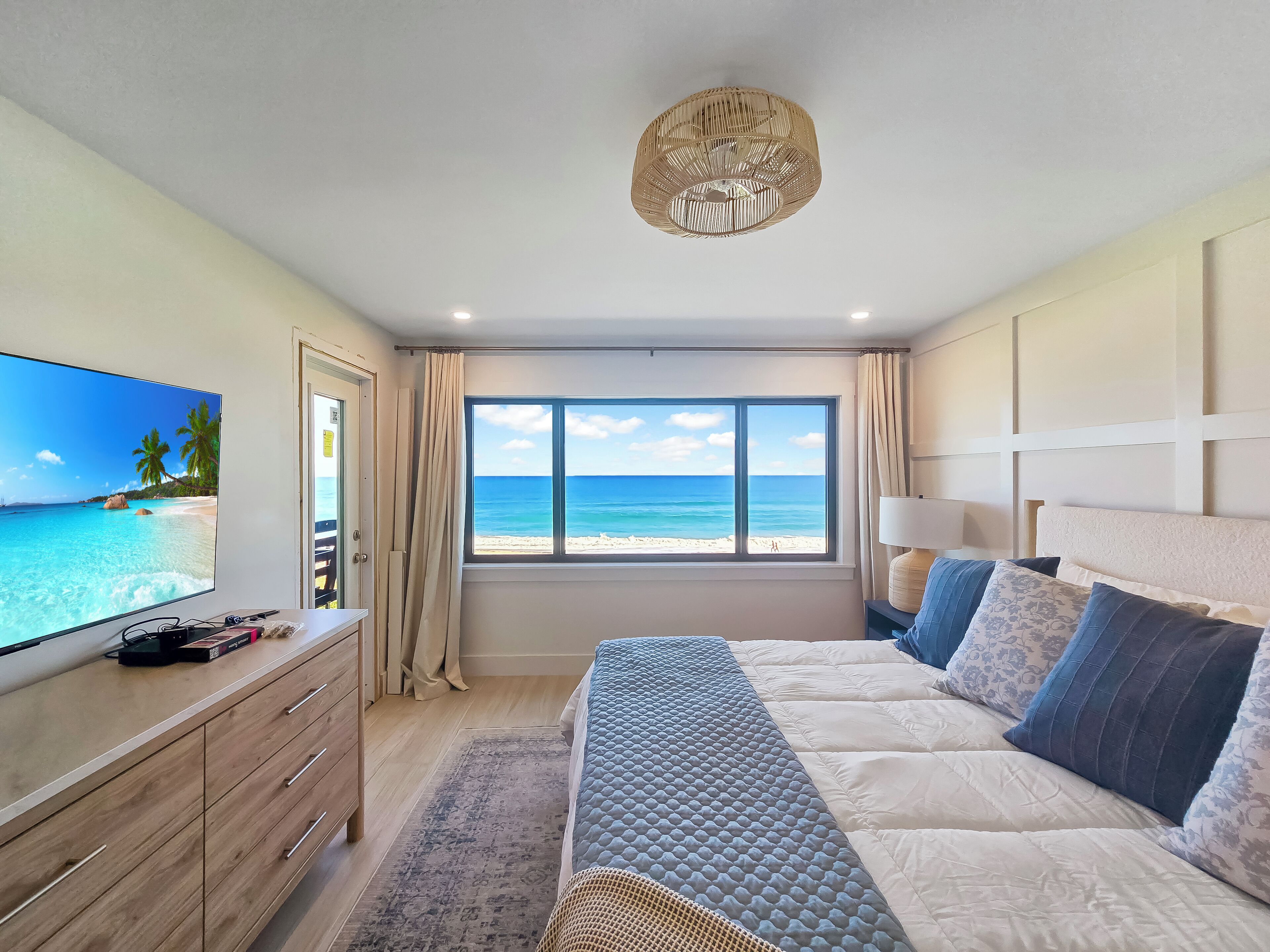 A bedroom with a large TV, a bed with multiple pillows, a window with a beach view, and a ceiling light.