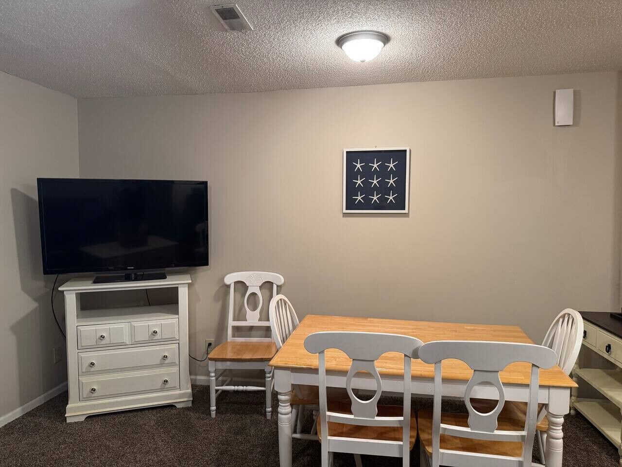 A room with a TV on a white stand, a wooden dining table with four chairs, and a wall-mounted picture frame.