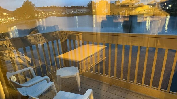 Een houten terras met een tafel en twee stoelen, met uitzicht op een waterpartij met weerspiegeling van gebouwen.