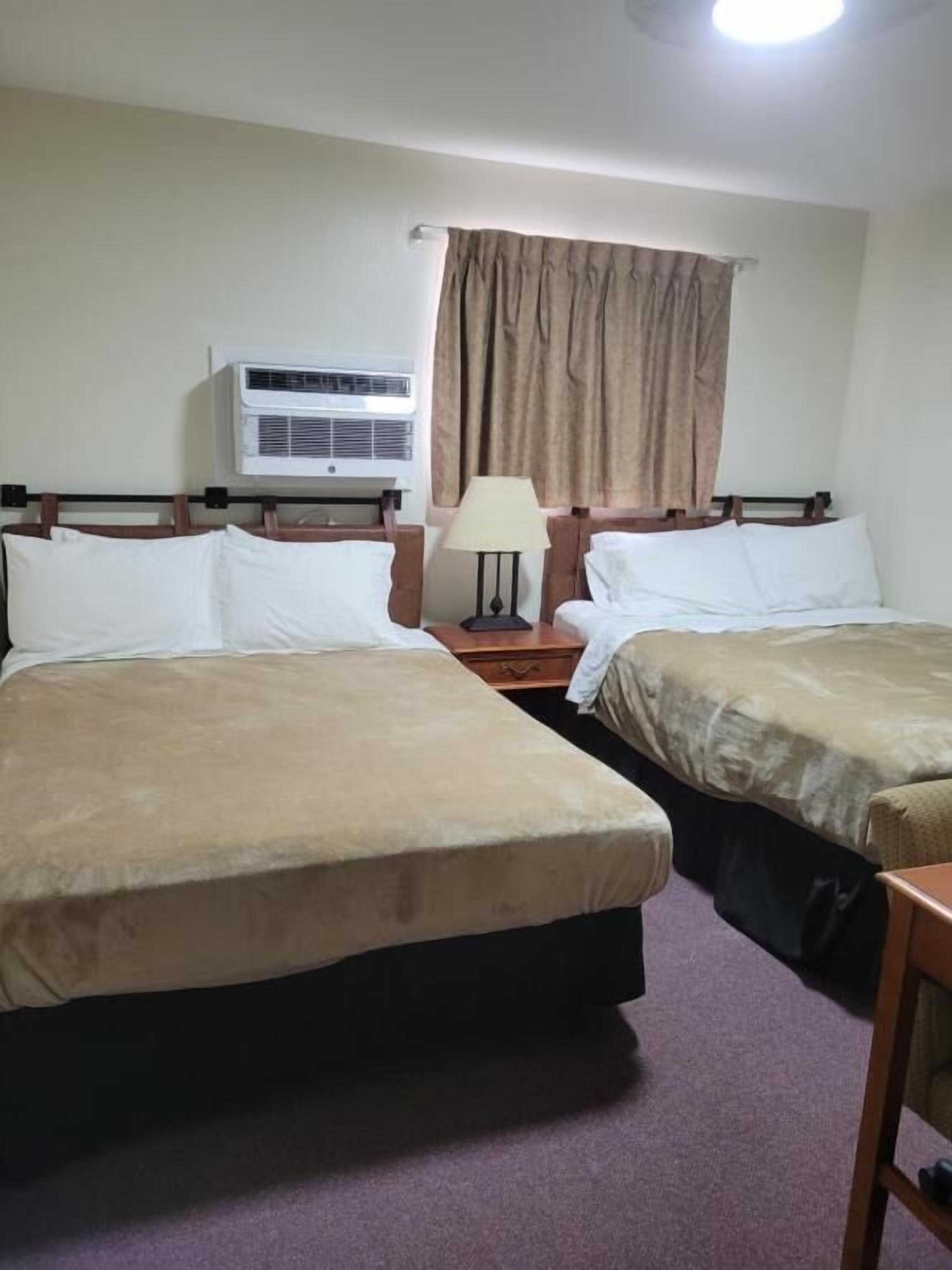 Habitación de hotel con dos camas, una mesita de noche con lámpara, un equipo de aire acondicionado y una ventana con cortinas.