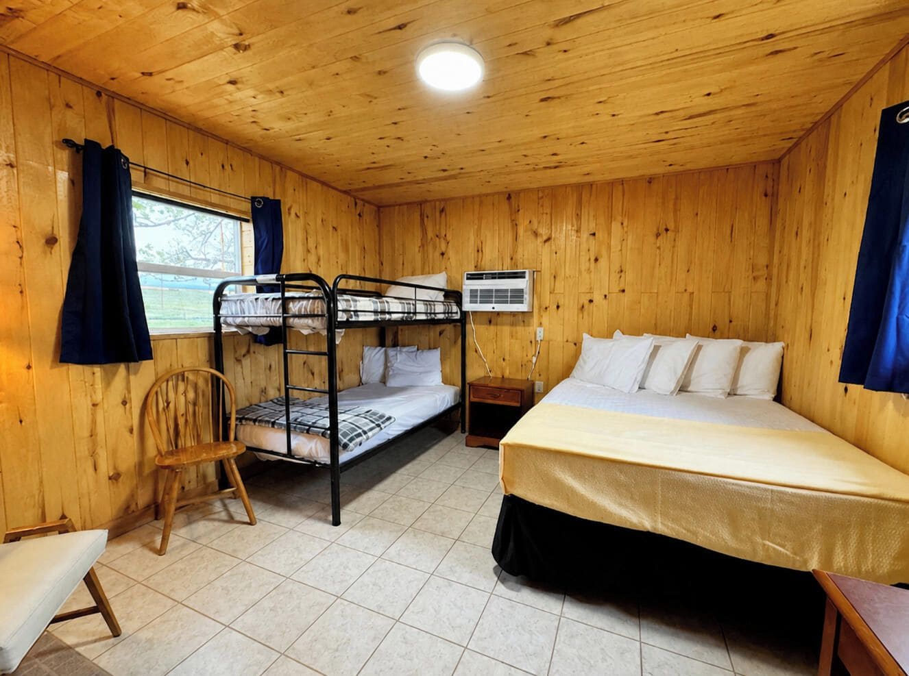 A room with wooden walls, a bunk bed, a single bed, a chair, a bedside table, and an air conditioning unit.
