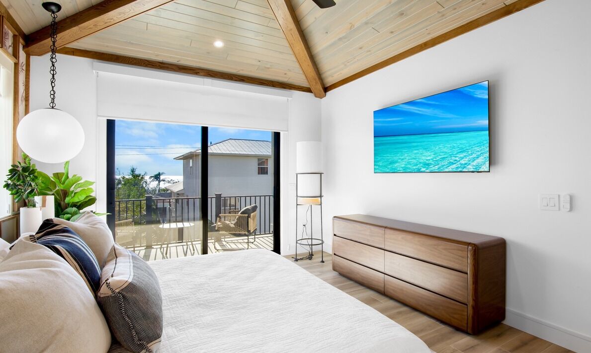 A bedroom with a large bed, a wooden dresser, a TV, and a balcony with a view of the ocean.